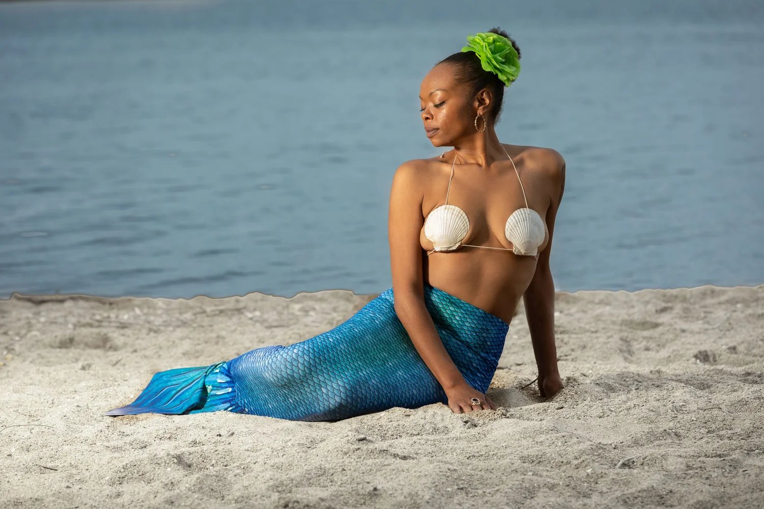Person dressed as a mermaid sitting on sandy beach with a blue mermaid tail and shell bikini top, evoking a touch of boudoir photography.